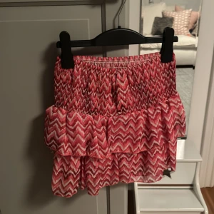 Rosa volangkjol med zickzackmönster - Supersöt kort kjol i rosa och vitt med zickzackmönster och volanger. Kjolen har smock i midjan för en bekväm passform och är perfekt för dig som gillar färg och mönster. Lätt och luftig, passar perfekt till varma dagar.