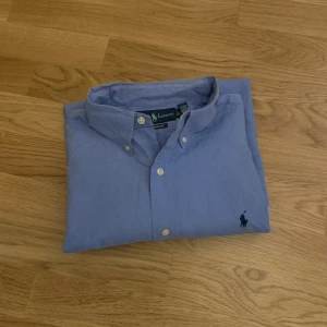 Blå skjorta från Ralph Lauren - Klassisk blå skjorta från Ralph Lauren med broderad logga på bröstet. Skjortan har lång ärm, button-down krage och vita knappar framtill. Modellen är Classic Fit och materialet är mjuk bomull. Perfekt för dig som gillar stilrena och tidlösa plagg.