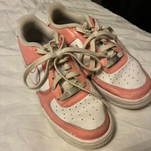 Nike Air Force 1 rosa och vita sneakers - Nike Air Force 1 sneakers i en snygg kombination av ljusrosa och vitt. Klassisk låg modell med rund tå, snörning och platt sula. Skorna har perforerade detaljer på tån och den ikoniska swoosh-loggan på sidan. Tillverkade i skinn med textilfoder för extra komfort.
