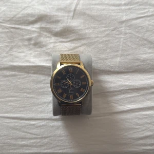 Guess guld klocka med mesh-armband - Snygg klocka från Guess med guldigt mesh-armband och svart urtavla. Urtavlan har romerska siffror i guld och tre mindre visartavlor. Klockan har en rund form och stilren design som passar till många outfits. Perfekt för dig som gillar klassisk men modern stil. 1 liten repa på glaset annars bra skick.