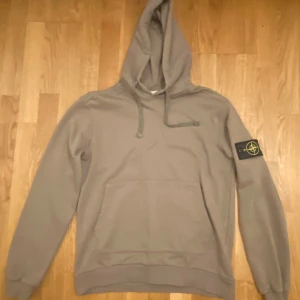 Mörk beige hoodie från Stone Island L - Tröjan sitter som en storlek M!     Säljer en hoodie från Stone Island med klassisk patch på vänster ärm. Tröjan har huva med snörning och en stor magficka framtill. Tillverkad i mjuk bomull och har en clean, stilren look som passar perfekt till streetwear.