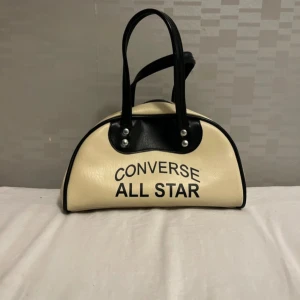 Converse All Star handväska i beige - Cool handväska från Converse All Star i beige och svart med tydlig logga framtill. Väskan har rundad form, svarta handtag och detaljer i silver. Materialet är syntet och den stängs med dragkedja med stjärndetalj. Perfekt för dig som gillar sportig streetstyle.