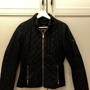 Svart quiltad jacka från Hollies - Quiltad svart jacka från Hollies med snygga guldfärgade dragkedjor och knappar. Insidan har ett klassiskt rutigt mönster i beige och svart. Jackan har lång ärm, ståkrage och figurnära passform. Perfekt för dig som gillar stilren och tidlös design.