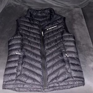 Svart dunväst från Peak Performance - Snygg svart dunväst från Peak Performance med quiltad design och dragkedja framtill. Västen har hög krage och två sidofickor med dragkedja. Perfekt för lager-på-lager och har en stilren look med diskret logga på bröstet.