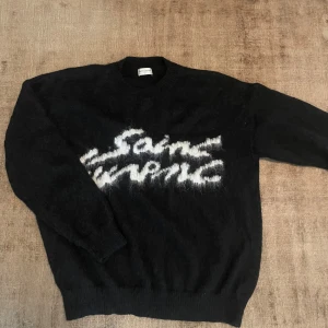Svart stickad Saint Laurent tröja stl S  - Svart stickad tröja från Saint Laurent med vit fuzzy logga framtill. Tröjan har rund halsringning, långa ärmar och ribbade muddar. Mjuk och lite luddig känsla, perfekt för kyliga dagar. Snygg och lyxig statement piece för din garderob. Tags medföljer. Kan gå ner lite i pris vid snabb affär. 