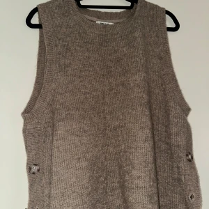 Beige stickad väst från Indiska - Stickad beige väst från Indiska med rund hals och snygga knappar på sidorna. Västen har en loose passform och är tillverkad i mjukt material som känns skönt mot huden. Perfekt att styla över en skjorta eller t-shirt för en chill look.