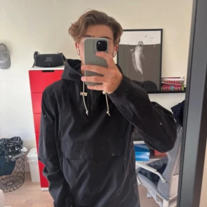 Svart vindjacka från The North Face - Säljer en svart vindjacka från The North Face i storlek M. Jackan har huva med snörning, half zip och en stor ficka framtill med lock. Snygg och enkel design med diskret logga på bröstet. Perfekt för dig som gillar streetwear och outdoor.