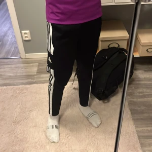 Svarta Adidas träningsbyxor med ränder - Svarta Adidas träningsbyxor med klassiska vita ränder längs sidorna och Adidas-logga på låret. Byxorna är i mjukt och stretchigt syntetmaterial, har resår i midjan och smal passform. Perfekta för sport eller chill. Primegreen-material.