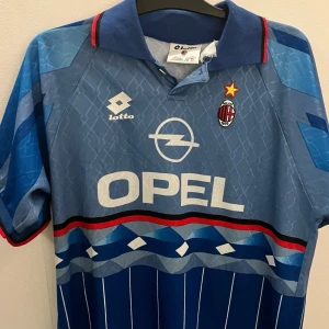AC Milan blå fotbollströja Weah #9 - Retro AC Milan fjärdetröja från säsongen 1995/96 med Weah #9 på ryggen. Tröjan är blå med vita och röda detaljer, har krage och korta ärmar. Snyggt mönster med ränder och geometriska former, samt klubbmärke och Opel-logga på bröstet. Perfekt för samlare eller Milan-fans.