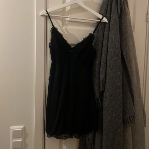 Svart spetsklänning med tunna axelband - Svart kortklänning med tunna justerbara axelband och v-ringad hals. Klänningen har fina spetsdetaljer både upptill och längs nederkanten, vilket ger en elegant och feminin look. Materialet är mjukt och följsamt, perfekt för dig som gillar stilrena och snygga plagg.