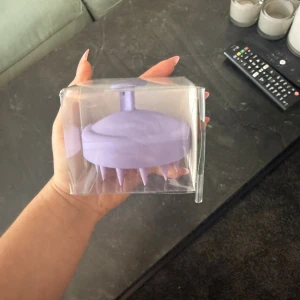 hårbottenmassageborste - Produkten på bilden är en lila hårbottenmassageborste i plast, förpackad i en genomskinlig ask. Den är avsedd för att stimulera hårbotten och förbättra blodcirkulationen. Produkten ser helt ny ut utan synligt slitage eller defekter.