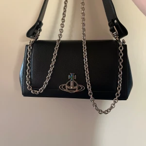 Svart axelväska från Vivienne Westwood - Snygg svart axelväska från Vivienne Westwood med kedjerem i silver och ikonisk metallogo framtill. Väskan är rektangulär och tillverkad i skinn med en stilren design som passar till många outfits.