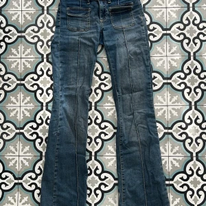 Blå bootcut jeans från Nelly - Snygga blå jeans från Nelly med bootcut passform och markerade sömmar längs benen. Klassiska fickor fram och bak, dubbla knappar i midjan och bälteshällor. Perfekta för dig som gillar en retroinspirerad look med lite utsvängda ben.