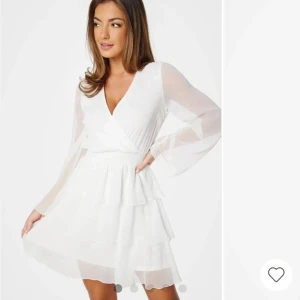 Vit omlottklänning med volanger storlek 36 - Säljer en vit kort omlottklänning med långa transparenta ärmar och v-ringad hals. Klänningen har flera lager volanger på kjolen och ett smockat midjeparti som ger en snygg siluett. Perfekt för dig som gillar romantisk och fräsch stil.