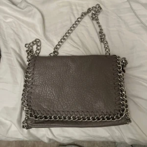 Grå axelväska med kedjedetaljer - Snygg grå axelväska i skinn med grov silverfärgad kedja som axelrem och runt kanterna. Väskan har en cool, lite ruff look och passar perfekt för dig som gillar statement-accessoarer. Perfekt storlek för det viktigaste och ger en edgy touch till din outfit.