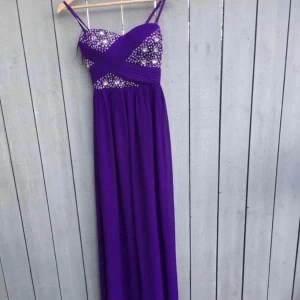 Lila långklänning med pärldetaljer - Långklänning i stark lila med smala axelband och v-ringad överdel. Livet är dekorerat med glittrande pärlor och stenar i ett mönster som ger extra wow-känsla. Kjolen faller rakt och mjukt från midjan. Perfekt för dig som vill sticka ut med färg och detaljer.passar storlek 34-36 jätte elegant resor per baksidan. Chiffong material stretch.
