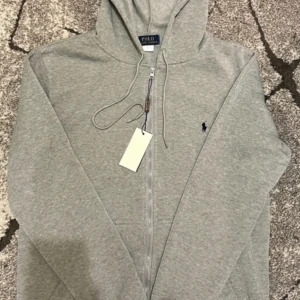 Grå hoodie från Polo Ralph Lauren - Snygg grå hoodie från Polo Ralph Lauren med dragkedja och huva. Tröjan har långa ärmar, snörning vid halsen och den klassiska Polo-loggan broderad på bröstet. Perfekt för chill dagar och enkel att matcha med jeans eller joggers.