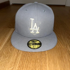 Grå LA Dodgers keps New Era 59FIFTY - Säljer en stilren grå LA Dodgers keps från New Era, modell 59FIFTY. Kepsen har broderad LA-logga framtill i vitt och MLB-logga bak. Tillverkad i 100% polyester med klassisk flat skärm och ventilerande hål upptill.