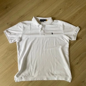 Polo Ralph Lauren - Vit pikétröja från Polo Ralph Lauren med klassisk krage och två knappar framtill. Tröjan har korta ärmar och den ikoniska broderade loggan i marinblått på bröstet. Tillverkad i mjuk bomull som är skön mot huden och passar perfekt till en clean och fräsch stil.