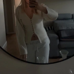 Vit v-ringad kofta med knappar - Säljer en vit v-ringad kofta med snygga knappar framtill. Koftan har långärmade ärmar och är croppad i modellen, vilket ger en modern och fräsch look. Materialet är mjukt och stickat, perfekt för att lyfta din outfit. Passar dig som gillar stilrena och trendiga plagg.