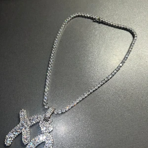 Glittrigt XO-halsband med stenar - Säljer ett blingat halsband med en stor XO-berlock täckt av gnistrande stenar. Kedjan är också dekorerad med glittrande stenar och har en silverfärgad finish. Perfekt för dig som vill sticka ut och addera extra shine till din look. Nypris 1300 kr. Oanvänd. 
