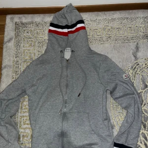 Grå Moncler hoodie med ränder - Snygg grå hoodie från Moncler med dragkedja och huva. Huva har coola ränder i svart, vit och rött. Tröjan har snörning vid huvan, fickor fram och Moncler-logga på ärmen. Perfekt för dig som gillar streetwear och vill ha något stilrent och bekvämt.
