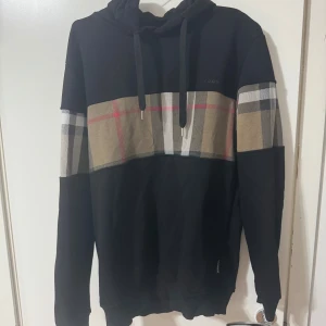 Svart hoodie med rutig Burberry-detalj - Svart hoodie från Burberry i storlek M med klassisk rutig panel i beige, röd, vit och svart över bröstet och ärmarna. Tröjan har huva med snörning och ribbade muddar. Perfekt för dig som gillar streetstyle och ikoniska mönster. Burrberry loggan är lite sliten man ser det i bilden annars är den fläckfri! 