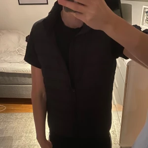 Svart quiltad dunväst med dragkedja - Säljer en svart dunväst med quiltad design och hög krage. Västen har en hel dragkedja framtill och är perfekt att slänga över en hoodie eller t-shirt.