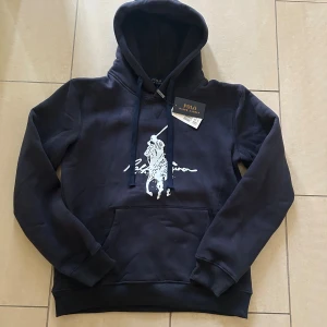 Marinblå hoodie från Polo Ralph Lauren - Svart hoodie från Polo Ralph Lauren med stor vit logga och tryck framtill. Klassisk känguruficka, snörning i huvan och mjukt material.  Färgen på tröjan kan skifta lite från bild till bild och kan se lite urtvättad,men färgen är så. Perfekt för dig som gillar stilren streetwear med premiumkänsla. Storlek L men passar även Storlek M