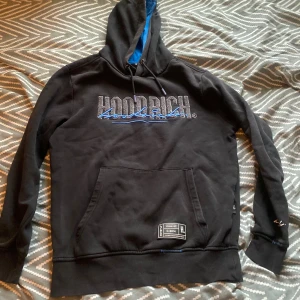 Svart hoodie från Hoodrich med blå detaljer - Svart hoodie från Hoodrich med broderad logga i grått och blått framtill och baktill. Fodrad huva med blå insida, känguruficka och snörning i halsen. Tillverkad i bomull och polyester, perfekt för dig som gillar streetwear.