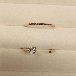 Två tunna guldringar med stenar - Säljer två stilrena guldfärgade ringar. Den ena är enkel och tunn med ett diskret mönster, medan den andra har en öppen design med en gnistrande klar sten och en liten detalj på motsatt sida. Perfekta för att mixa och matcha för en trendig look.