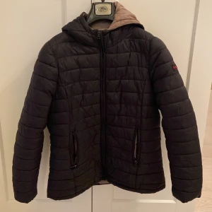 Svart pufferjacka från Marikoo - Svart pufferjacka från Marikoo med quiltad design och huva. Jackan har dragkedja framtill och två fickor med dragkedja. Insidan är beige och jackan har en broderad logga på ärmen. 