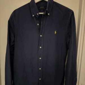 Mörkblå skjorta Polo Ralph Lauren - Slim fit mörkblå skjorta från Polo Ralph Lauren med klassisk krage och knappar framtill. Broderad gul logga på bröstet. Skjortan har långa ärmar och är tillverkad i mjuk bomull som känns skön mot huden. Perfekt för dig som gillar stilrena och tidlösa plagg.