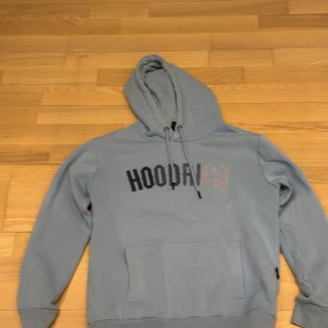 Blå hoodie från Hoodrich med broderi - Snygg ljusblå hoodie från Hoodrich i storlek S. Tröjan har broderad logga i mörkblått och ljusrosa på bröstet, snörning vid huvan och en stor magficka. Tillverkad i mjuk bomullsmix, perfekt för chill eller streetwear. Huvan är rymlig och ärmarna är långa.