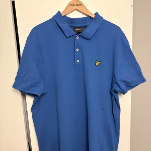 Blå pikétröja från Lyle & Scott xxl - Säljer en klassisk blå pikétröja från Lyle & Scott med gul logga på bröstet. Tröjan har krage, tre knappar framtill och korta ärmar. Perfekt för dig som gillar stilrena och sportiga plagg. Materialet är mjuk bomull.