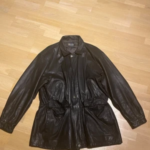 Svart skinnjacka från Molltan - Svart skinnjacka från Molltan med klassisk krage, stora fickor med lock och knappar samt justerbart midjebälte. Jackan har lång ärm och en loose passform, perfekt för dig som gillar retrovibe och vill ha en cool look. Fodrad insida för extra komfort.