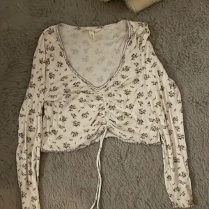Blommig croppad topp från H&M - Säljer en ljuslila och vit croppad topp från H&M med blommigt mönster och ribbad struktur. Toppen har lång ärm, v-ringning med dekorativ söm och snörning framtill som ger en snygg rynkad effekt. Perfekt för dig som gillar romantisk och trendig stil. Den är storlek M men passar XS/S