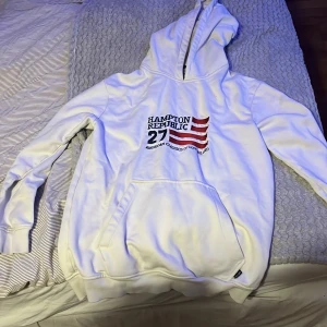 Vit hoodie från Hampton Republic - Vit hoodie från Hampton Republic med tryck framtill i blått och rött samt amerikansk flaggdetalj. Klassisk känguruficka och huva. Perfekt för en chill och sportig look.