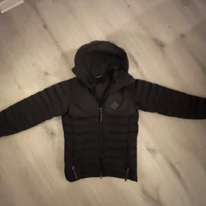 Svart dunjacka från J.Lindeberg - Svart dunjacka från J.Lindeberg med huva och hel dragkedja framtill. Jackan har två sidofickor med dragkedja och ett diskret märke på bröstet. Den är quiltad och har en clean, sportig look som passar perfekt när det är kallt ute.