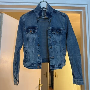 Blå jeansjacka från &Denim, strl 34 - Snygg klassisk blå jeansjacka från &Denim i storlek 34. Jackan har två bröstfickor med knappar, framfickor och knappar längs hela framsidan. Långärmad med krage. 