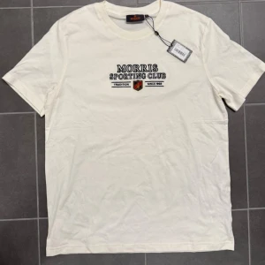 Offwhite Morris Sporting Club t-shirt - Snygg offwhite t-shirt från Morris Sporting Club med broderad logga och emblem på bröstet. Klassisk rund hals och korta ärmar. Tillverkad i mjuk bomull för skön känsla. Perfekt för dig som gillar stilrena och sportiga plagg med en twist.