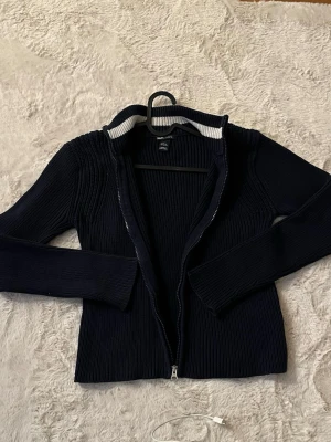 Marinblå ribbad kofta från H&M L.O.G.G. - Säljer en marinblå ribbstickad kofta från H&M L.O.G.G. med dragkedja framtill och vit rand på insidan av kragen. Långärmad och i mjukt bomullsmaterial, perfekt för lager på lager under kyliga dagar. Passar S