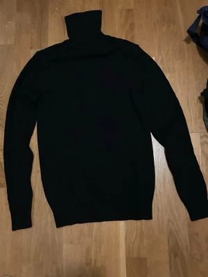 Svart polotröja i stickat material - En svart polotröja i stickat material med lång ärm och hög krage. Perfekt för kyliga dagar och enkel att matcha med olika outfits. Klassisk och stilren design som funkar till det mesta.