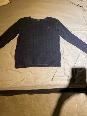 Mörkblå kabelstickad tröja Polo Ralph Lauren - Kabelstickad mörkblå tröja från Polo Ralph Lauren med klassisk röd logga broderad på bröstet. Tröjan har rund halsringning, ribbade muddar och är långärmad. Perfekt för dig som gillar stilren..