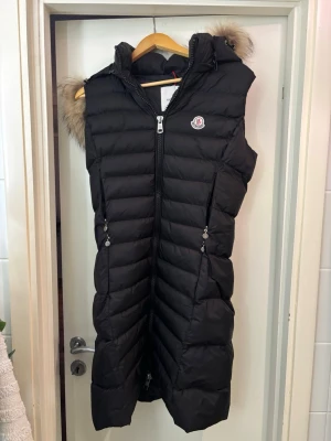 Svart lång dunväst från Moncler - Säljer en svart lång dunväst från Moncler med huva och snygg pälsdetalj. Västen har dragkedja framtill, två fickor med dragkedja och klassisk Moncler-logga på bröstet. Perfekt för kyliga dagar när du vill vara både varm och stilren. Västen är i strl S, men passar även en M