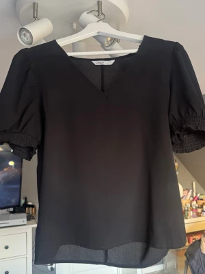 Svart blus med puffärm från ONLY - Säljer en svart blus från ONLY med v-ringning och puffiga korta ärmar med smockdetalj. Blusen har en lätt och luftig känsla, perfekt till många olika looks. Enkel men ändå stylish design som är lätt att matcha.