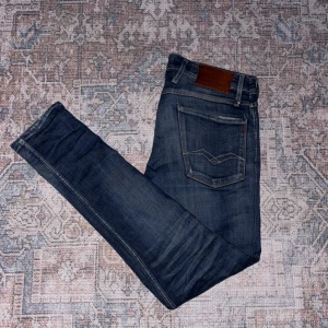 Replay Anbass jeans mörkblå slim - Mörkblå Replay Anbass jeans med snygg tvätt och klassiska kontrastsömmar. Modellen har fem fickor, läderpatch bak och smal passform. Jeansen är tillverkade i stretchig bomullsdenim för extra komfort och har diskreta slitningar på låren.