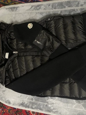 Svart Moncler pufferjacka med stickade  ärmar Size Xl - Säljer en svart pufferjacka från Moncler med stickade ärmar och rygg. Jackan har dragkedja framtill, ficka med dragkedja på ärmen och klassisk Moncler-logga. Kombinerar glansigt quiltat material med ribbstickade partier för en modern look.