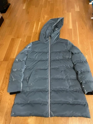 Blå lång pufferjacka från Dolcezza - Säljer en lång, blå pufferjacka från Dolcezza med huva och dragkedja framtill. Jackan har en clean look med två sidofickor och quiltad design. Perfekt för kalla dagar och riktigt snygg till både jeans och boots.