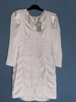 Beige kort klänning från H&M - Säljer en beige kort klänning från H&M i storlek 38. Klänningen har v-ringning, långa ärmar och ett snyggt rynkat tyg framtill som ger en cool struktur. Perfekt för dig som vill ha en stilren men ändå unik look.